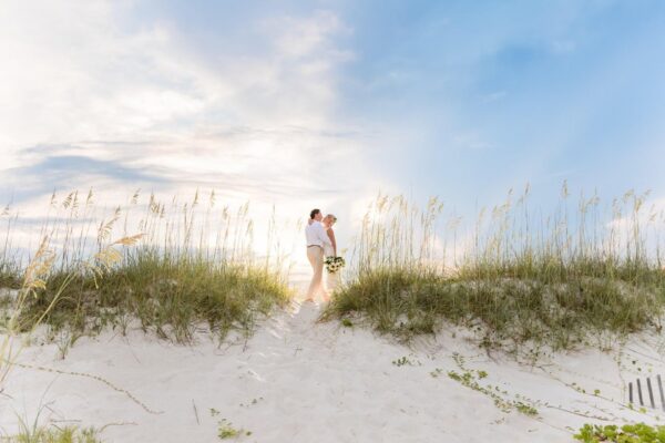 Two people dressed in white stand together on a sandy beach dune with tall grass, holding flowers, under a partly cloudy sky with sunlight streaming through.