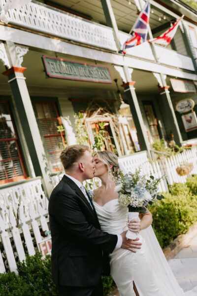 A bride and groom kiss in front of a historic inn with white railings and flags. The bride holds a bouquet and wears a white dress; the groom wears a black suit.