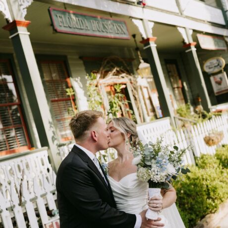 A bride and groom kiss in front of a historic inn with white railings and flags. The bride holds a bouquet and wears a white dress; the groom wears a black suit.