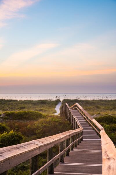 Wooden boardwalk leading through green dunes toward a beach, with calm ocean and a colorful sunrise or sunset sky in the background.
