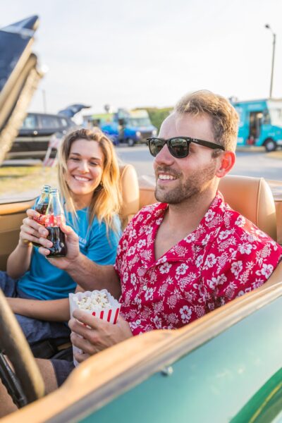 A smiling man in a red floral shirt holds popcorn and a woman holds a soda bottle as they sit in a convertible car, enjoying a sunny day at an outdoor event.