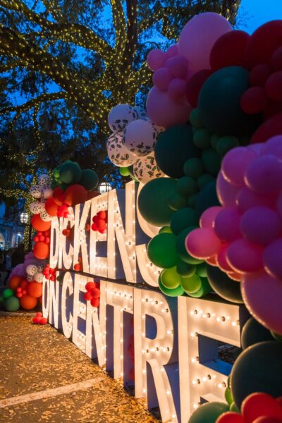 Large marquee letters lit up read DICKENS ON CENTRE, surrounded by colorful red, green, pink, and white balloon decorations, with string lights in trees above during an outdoor evening event.