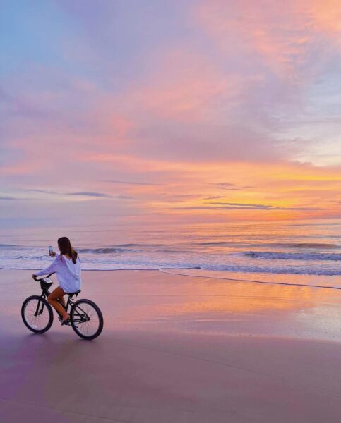 A person rides a bicycle along a sandy beach at sunrise or sunset, holding up a phone to take a photo. The sky is filled with pastel colors, reflected on the calm ocean and wet sand.