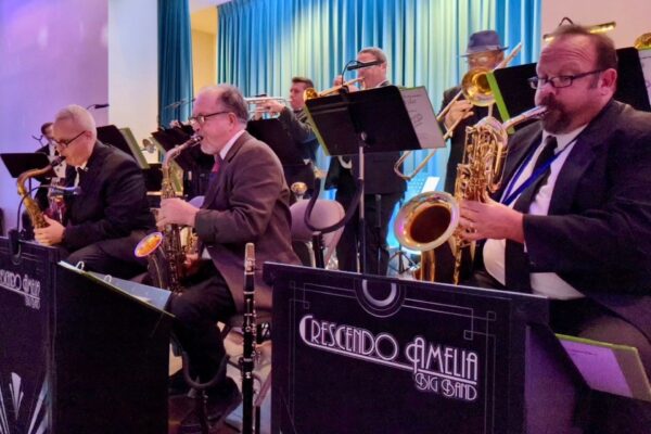 A jazz big band called Crescendo Amelia performs on stage, with musicians playing saxophones, trumpets, and trombones in front of blue curtains. The band members are dressed formally in suits.