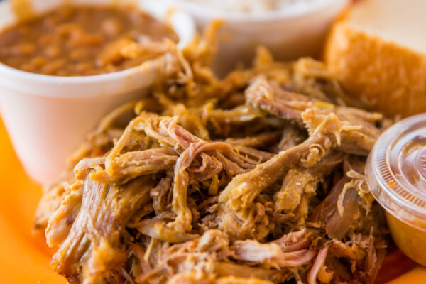 Close-up of pulled pork served on a plate with baked beans, a slice of bread, and a small container of sauce in the background. The food is arranged in a casual, appetizing manner.