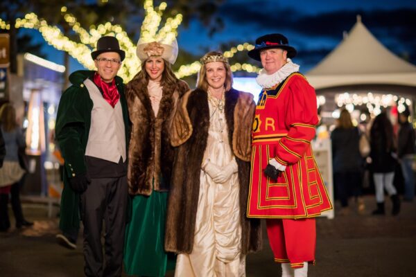 Four people dressed in festive Victorian-era costumes stand together and smile at an outdoor event, with string lights and a white tent glowing in the background at dusk.