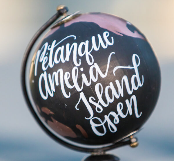 A small black globe with white cursive text reading Pétanque Amelia Island Open is displayed, with blurred background.