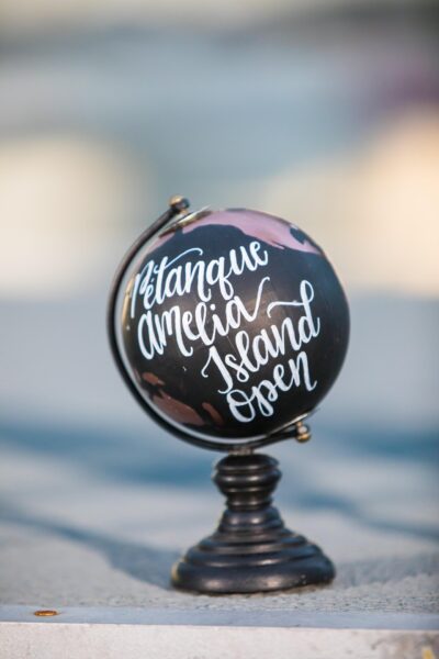 A small black globe on a stand with white cursive writing that reads Pétanque Amelia Island Open, set against a blurred background.