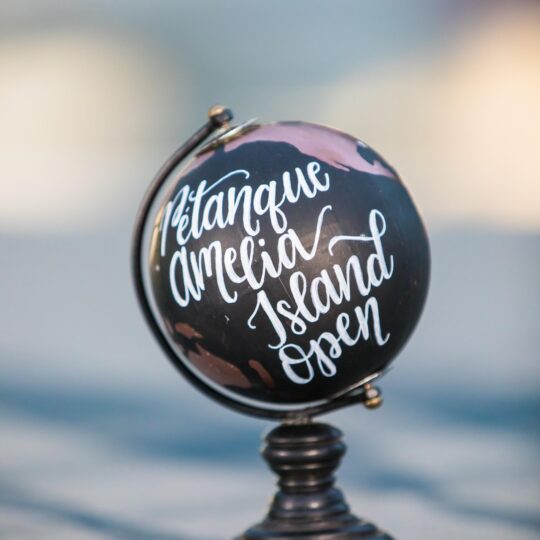 A small black globe on a stand with white cursive writing that reads Pétanque Amelia Island Open, set against a blurred background.