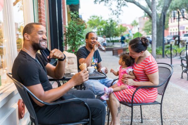 Three adults and a toddler sit at an outdoor café table. Two men are smiling and eating ice cream, while a woman, in a striped dress, holds and kisses the toddler. Trees and shops are visible in the background.