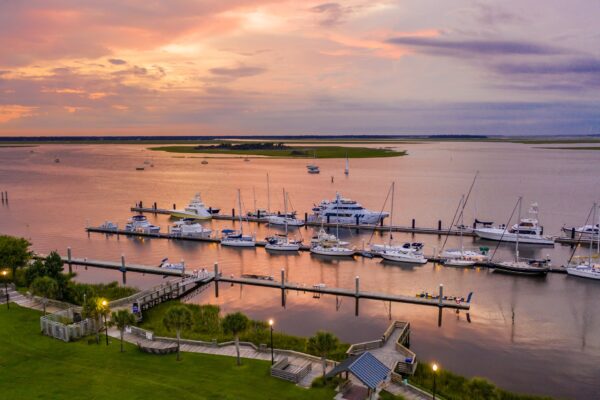 A marina at sunset with yachts and boats docked along several piers, calm water reflecting the colorful sky, and green grassy areas in the foreground.