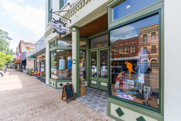 A clothing boutique with green trim and large windows displays summer outfits, hats, and accessories. Across the street, red brick buildings are visible. A sandwich board sits on the sidewalk.
