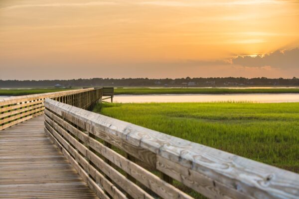 A wooden boardwalk curves through green marshland toward a distant viewing platform, with a calm river and orange sunset sky in the background.