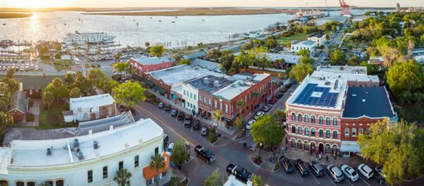 Aerial view of a coastal town at sunset, showing brick buildings, tree-lined streets, parked cars, and a waterfront with boats and docks in the background.