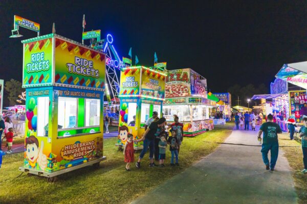 A brightly lit carnival at night features ticket booths, a pizza stand, and various rides in the background. People, including families with children, walk and gather along the pathway.