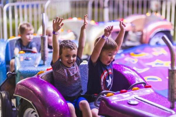 Two young boys sit in a bright purple car on a carnival ride with their arms raised, smiling and laughing, while other children ride in cars behind them. The background features colorful lights and a blurred fence.