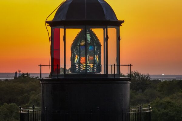 A lighthouse lantern glows brightly at sunset, with its glass Fresnel lens reflecting colorful light. The sky is orange and yellow, and green trees are visible below the lighthouse.