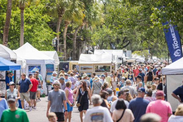 A large crowd walks through an outdoor art festival lined with white tents and booths on a sunny day. People of all ages browse artwork and vendors, surrounded by green trees and festive atmosphere.