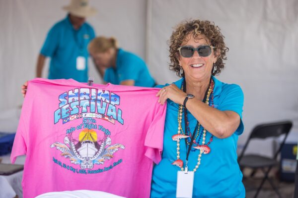 A smiling woman wearing sunglasses holds up a bright pink Shrimp Festival T-shirt. She stands at a festival booth with two other people in blue shirts blurred in the background.
