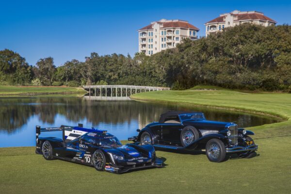 Two cars, a modern race car and a vintage convertible, are parked side by side on a golf course near a pond, with trees, a footbridge, and multi-story buildings in the background under a clear blue sky.