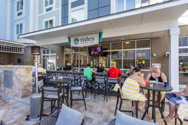 People relax and enjoy drinks at an outdoor poolside bar called Tides Pool Bar & Grill, located at a modern hotel. Some sit at the bar, others at tables, with a TV above the bar and sunny weather outside.