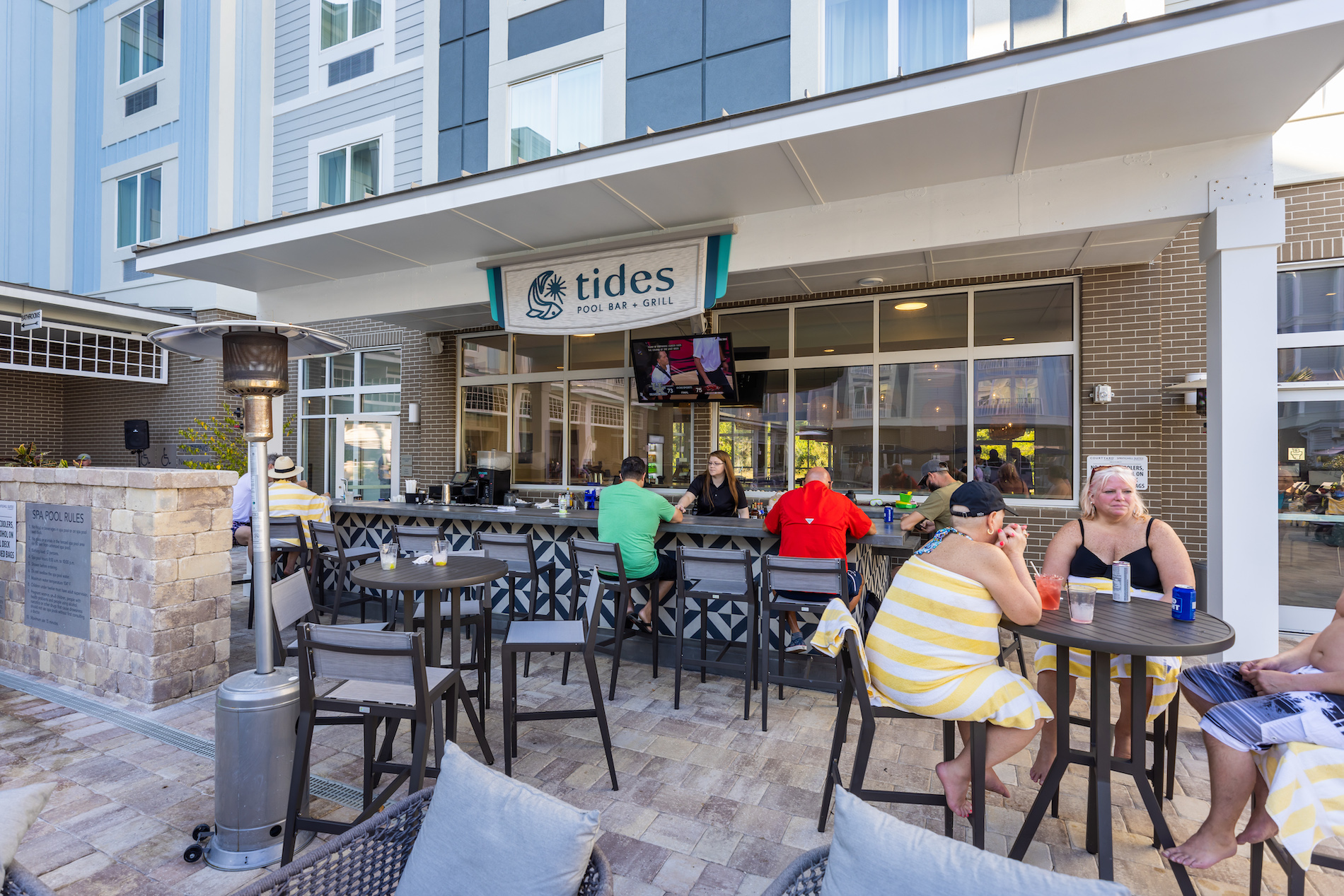 People relax and enjoy drinks at an outdoor poolside bar called Tides Pool Bar & Grill, located at a modern hotel. Some sit at the bar, others at tables, with a TV above the bar and sunny weather outside.