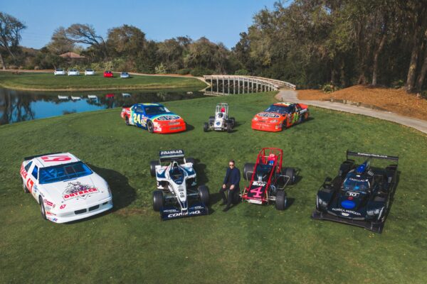 A man stands on a grassy lawn beside seven different race cars arranged in a semicircle, near a pond and a small arched bridge, with trees in the background under a clear sky.