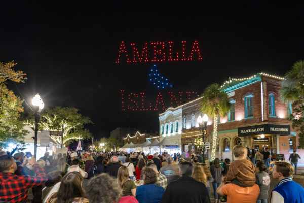 A nighttime street scene with a crowd watching drones form the words “Amelia Island” in red lights above historic brick buildings decorated with lights and palm trees.