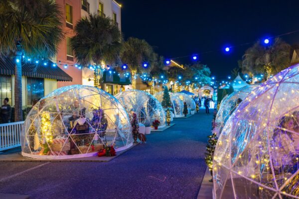 A festive street at night is decorated with string lights and features clear, igloo-shaped domes filled with tables, chairs, and holiday decorations, creating cozy outdoor dining spaces.