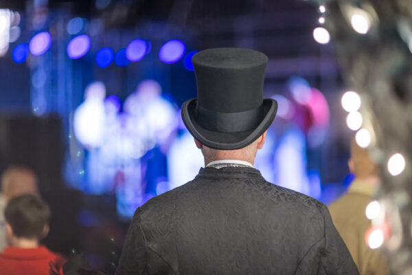 A person in a black top hat and ornate coat stands with their back to the camera, watching a brightly lit stage show with blurred figures and blue lights in the background.