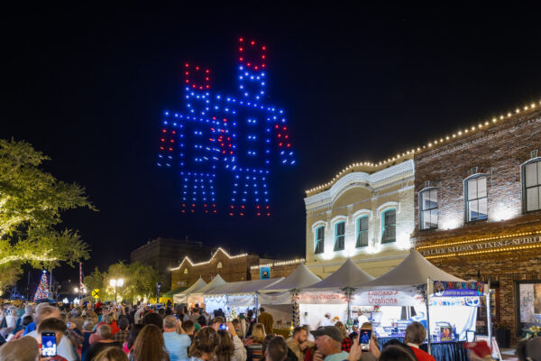 A crowd watches as hundreds of drones form two large, colorful robots in the night sky above a festive street with market stalls and illuminated historic buildings.