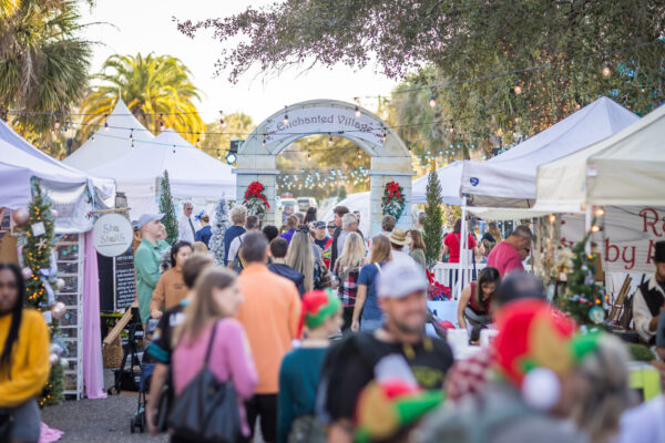 A festive outdoor market with people walking among white tents decorated with holiday ornaments, lights, and garlands. An archway labeled Enchanted Village is visible in the background under palm trees.