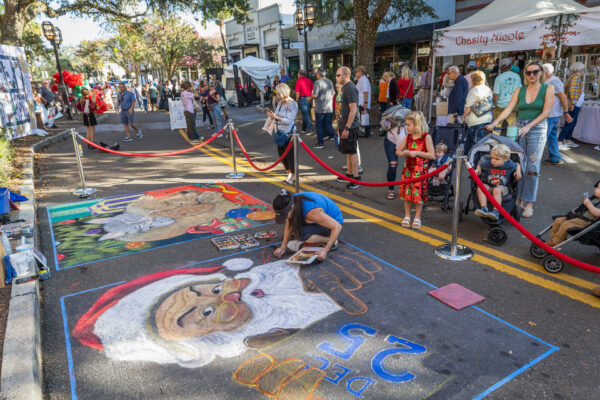 A street artist draws a colorful Santa Claus chalk mural on pavement at an outdoor festival, with people watching behind red ropes and vendor tents lining the street on a sunny day.