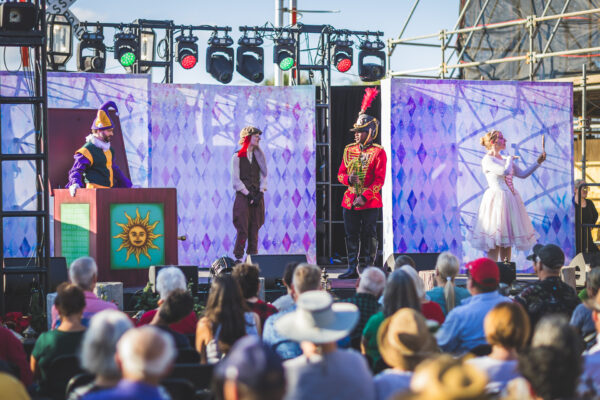A costumed theater performance takes place on an outdoor stage with four actors in colorful outfits, while an audience sits and watches under a sunny sky.