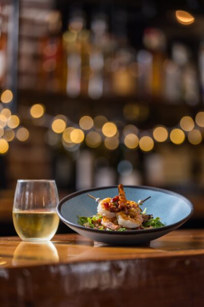 A glass of white wine sits next to a plate of grilled shrimp skewers on a bed of greens, with a softly lit bar and string lights blurred in the background.