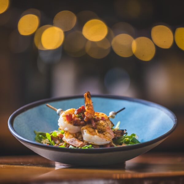 A close-up of shrimp skewers garnished with diced vegetables, served on a bed of greens in a blue bowl, with warm bokeh lights blurred in the background.