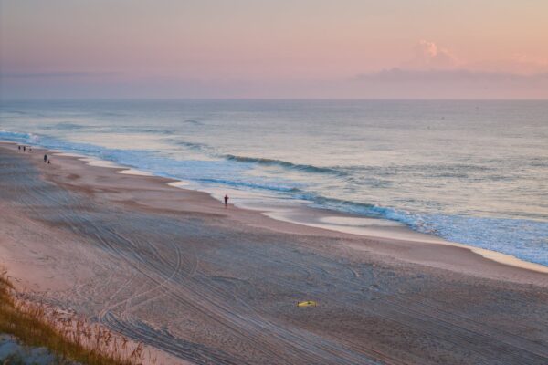 A tranquil beach at sunrise with gentle waves, soft pastel skies, and a few distant people walking along the shore. Tire tracks and footprints are visible in the sand with sparse vegetation in the foreground.