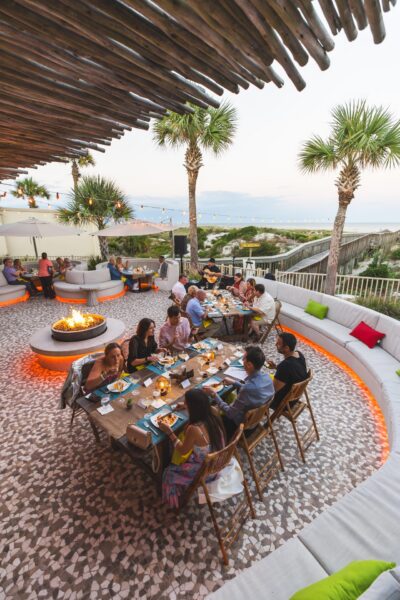 A group of people dine at a long outdoor table near a fire pit, surrounded by palm trees and ambient lighting, with a view of the ocean in the background.