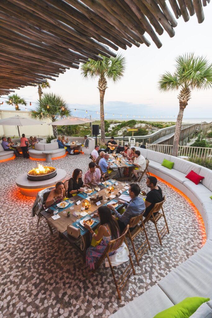 A group of people dine at a long outdoor table near a fire pit, surrounded by palm trees and ambient lighting, with a view of the ocean in the background.