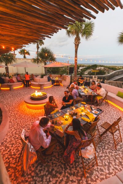 People dining at an outdoor restaurant with warm orange lighting, a central fire pit, palm trees, string lights, and a view overlooking the ocean at sunset. The atmosphere is cozy and relaxed.
