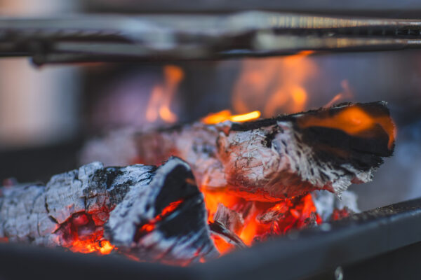 Close-up of glowing, burning logs with bright orange embers and ash in a fire pit, with metal grill bars visible above the flames.