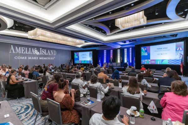 A large conference room filled with seated attendees facing a stage, where a speaker presents slides. The backdrop reads “AMELIA ISLAND COME MAKE MEMORIES!” The room is well-lit with modern chandeliers.