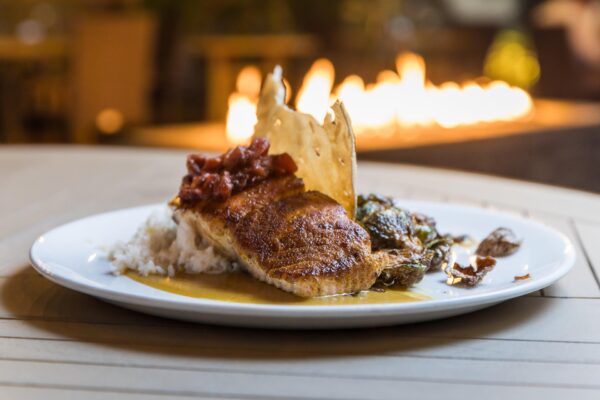 A plated dish featuring a seasoned, grilled fish fillet atop white rice, garnished with a crispy cracker, roasted vegetables, and a tomato-based sauce, with a blurred fire in the background.