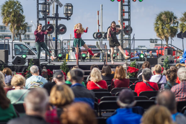 Four performers sing and dance on an outdoor stage, entertaining a seated audience. The scene is festive with holiday decorations, and palm trees are visible in the background under a clear sky.