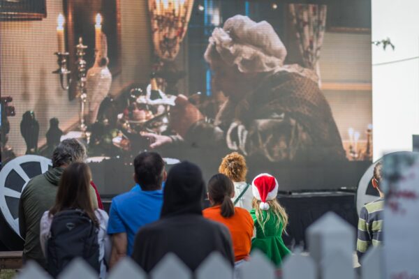 A group of people, including a child in a Santa hat, watch an outdoor screening of a classic film showing a person in old-fashioned attire eating at a candlelit table.