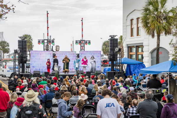 A crowd watches performers in colorful costumes on an outdoor stage at a waterfront event, with palm trees, tents, and a large building in the background.