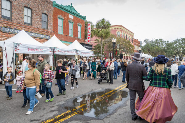 A busy outdoor street festival with white vendor tents, people walking, and two individuals in Victorian-style costumes. Historic brick buildings and a theater sign reading PALACE are visible in the background. A puddle reflects the scene.