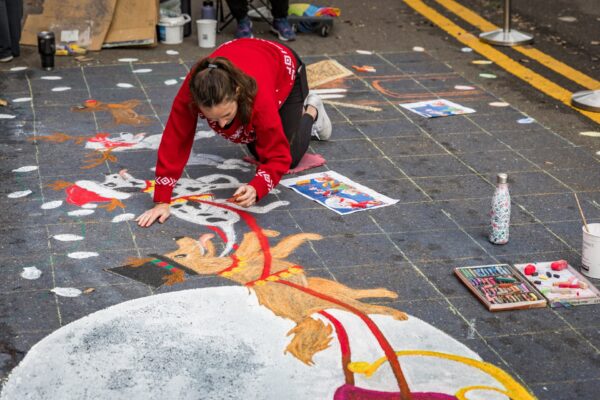 A woman in a red sweater kneels on a street, creating a colorful chalk drawing of reindeer and snow on the pavement. Art supplies and reference images are scattered around her.