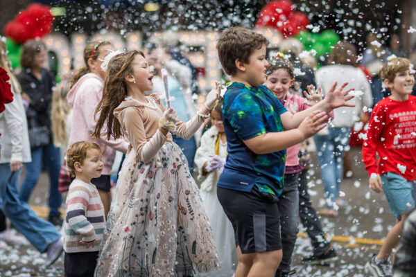 Children joyfully play in artificial snow at an outdoor event, with a girl in a floral dress smiling and holding a wand, surrounded by kids in colorful clothing and festive decorations in the background.