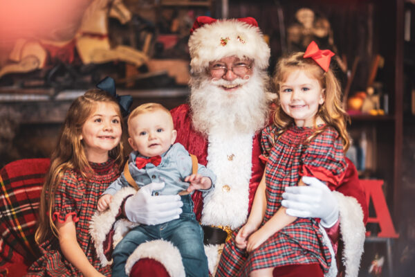 Santa Claus sits in the center, smiling, with two young girls in plaid dresses and bows, and a baby boy on his lap, all smiling. The background has festive holiday decorations.